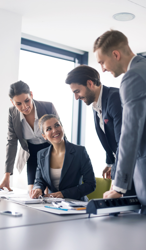 Four business professionals in a meeting, gathered around a table with documents, discussing their new managed Keycloak service strategy. Three are standing while one is seated, all engaged in the vibrant office setting. managed keycloak service, keycloak managed, managed keycloak, managed keycloak hosting