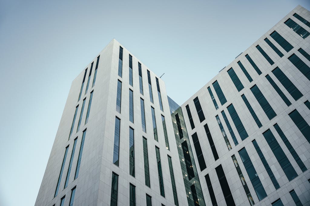 From below of contemporary multistory building with futuristic design and windows with unusual shapes located against cloudless blue sky on street of city